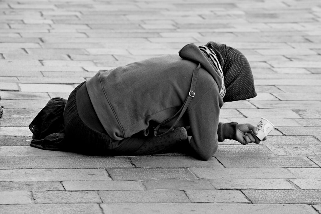 Schwarz-Weiß-Aufnahme einer bettelnden Person in einer venezianischen Gasse, die tief gebeugt am Boden kniet.